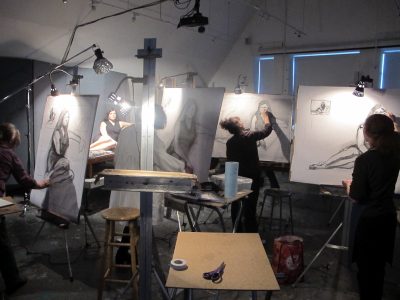 Darkened art studio, with multiple people drawing in charcoal on large easels from a life model.
