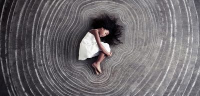 Dark skinned child in a white dress lays on a black chalkboard background, with white chalk lines traced around her form radiating outward.