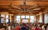Crowded event space with a rustic wood interior. A large fan is installed from the ceiling. A projector screen in the front of the room displays an ongoing presentation.