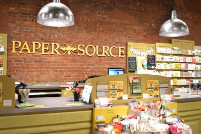 Retail store interior with a display shelf of cards and paper goods in the foreground and background. Sign on the wall reads "Paper Source"
