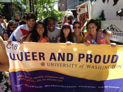 Group of students hold a banner in a parade line up, reading "Queer And Proud @ University of Washington"