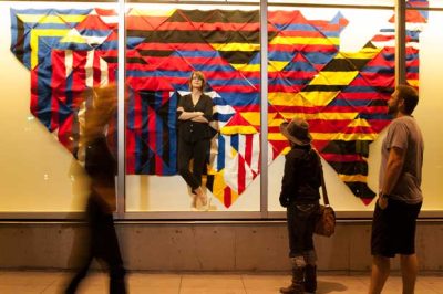 Multicolored art installation of 2D square shapes in a large storefront window. A person stands inside, and another outside looking in.