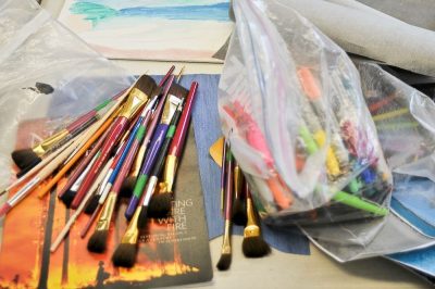 Art supplies laid out over a desk; there's a bundle of paintbrushes, a ziploc of shapries and pens, on tops of some canvases and paper collages.