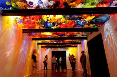 Museum visitors stand underneath a ceiling art installation featuring hundreds of small colorful organic glass pieces, lit from behind so they case colorful shadows