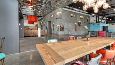 Modern open floor plan office space with exposed concrete floor and walls. Large polished wood eating table in the foreground with several brightly colored chairs around it.