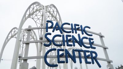 Blue metal lettering reads "Pacific Science Center" mounted on a white metal sign. White archways rise in the distance behind the sign against a gray sky.