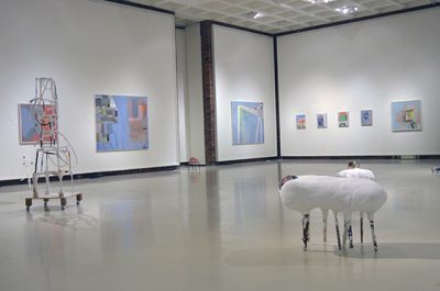 Empty, brightly lit gallery with pastel abstract paintings hung on the wall and abstract sculptures in the center of the space.