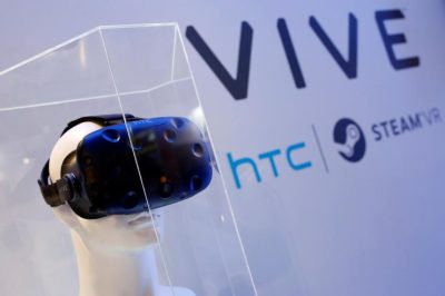 Virtual reality headset is displayed on a mannequin head in a clear vitrine. In the background, a sign reads "Vive; HTC; Steam VR"
