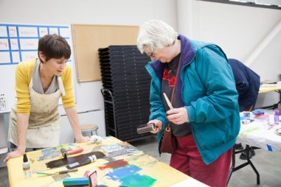 Two people in a studio setting working at a printmaking stations. One holds a brayer with ink while the other leans over the table.