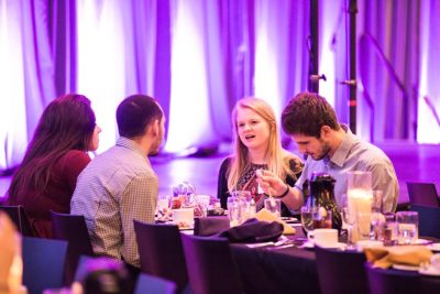 Group of 4 students chat at a reception lunch gathering, a purple lit stage in the background.