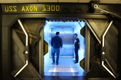 Futuristic space with sliding metal doors and the text "USS Axon 1300" above. Two people stand in an airlock beyond the entrance.