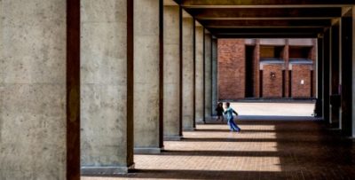 Breezeway of a university building featuring striking concrete columns and red brick ground. A child runs between the columns.