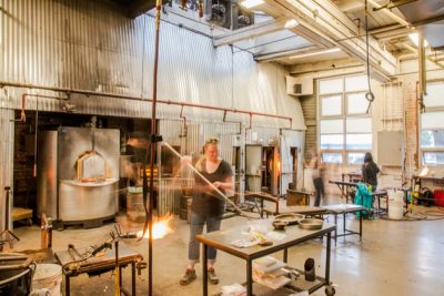 Brightly lit glassblowing studio with people working at different stations. Person in the foreground works with a blowpipe molding glass on a table.