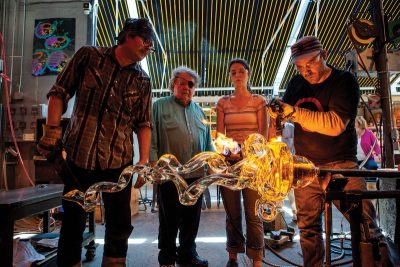 Group of four people stand around a blown glass art piece on the end of a blow pipe in a glassblowing studio. One person holds a blow torch to the piece of glass.