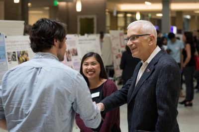 Two people shake hands in the midst of a research fair, while a third person smiles behind them.
