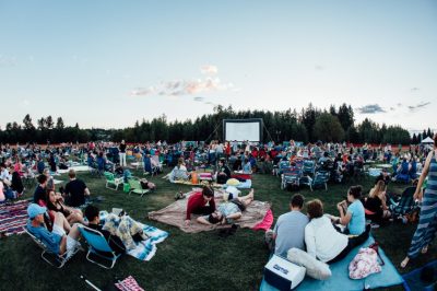 People sitting on blankets and in chairs on a large lawn for an outdoor movie screening.