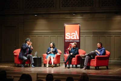 Group of four people on a stage seated in red chairs, a banner reading "SAL: Seattle Arts & Lectures" on stage