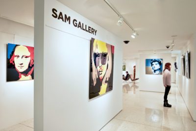Bright, white walled gallery with painting hung on the wall. Wall text reads "SAM Gallery." A person looks at one of the artworks on display.