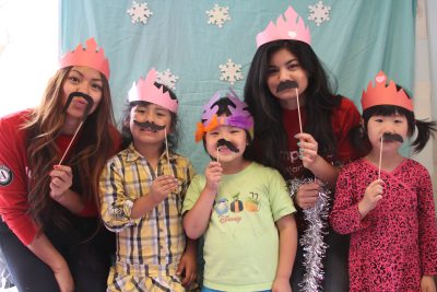Two college students and three young children stand in front of a light blue backdrop, wearing construction paper crowns and mustaches.