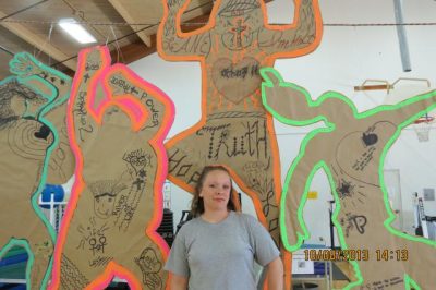 Person stands in front of large brown paper silhouette artworks of human figures, with text and images drawn inside.