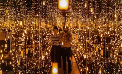 Two people stand inside an art installation by artist Yayoi Kusama consisting of an entirely mirrored room and reflected lights