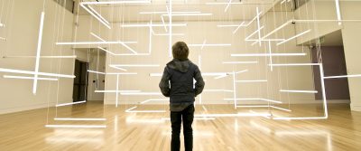 Person stands in an art gallery in front of an installation piece featuring tube lightbulbs