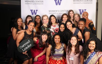 Group of femme people stand in front of a photo backdrop with purple "W"s and "Women's Center - University of Washington" imprinted on it. One holds a sign reading "Current Leader"