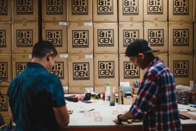 Two people work at a studio production table, a wall of cardboard boxes behind them with company logos on the outside that read "8th Gen"