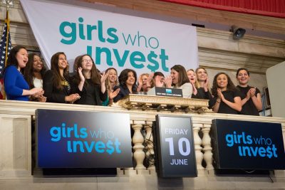Group of applauding people standing in front of a banner that reads "Girls Who Invest"