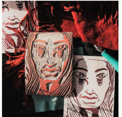 A linocut block of a portrait, with completed prints and carving tool to the left and right of it.
