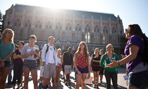 Group of students stand in front of a multi-storied, historical University building, listening to a tour guide in front of the group.
