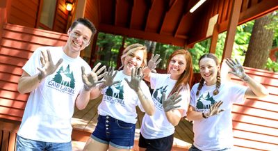 Group of 4 smiling young adults in matching summer camp t-shirts hold up clay covered hands.
