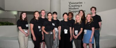 Group of smiling young adults standing in front of a museum exhibition title wall that reads "Visionaries: Creating a Modern Guggenheim"
