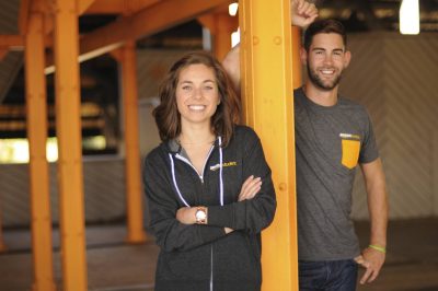 Two smiling young adults stand in an open floor plan office space, both wearing "Amazon Student" branded apparel.