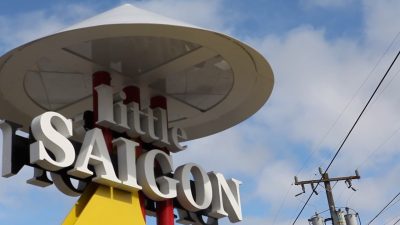 Large metal street sign that reads "Little Saigon" in white metal letters against a blue and white clouded sky.