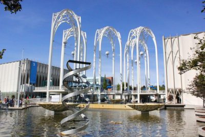 Five white modern arches stand between two large museum buildings, a fountain in the foreground featuring a metal public art sculpture.
