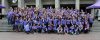 Large group of college aged youths gather over a series of stone steps, all gesturing and smiling at the camera. They are all wearing matching purple tshirts.