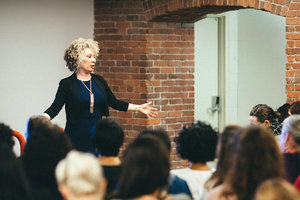 Femme person stands in front of an audience giving a talk, hands outspread.