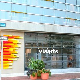 Windowed building exterior with white vinyl lettering that reads "visArts"; a red, orange and yellow wall art installation is to the left.