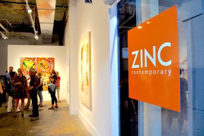 White walled gallery with groups of visitors looking at art and chatting; orange door sign reads "Zinc Contemporary"