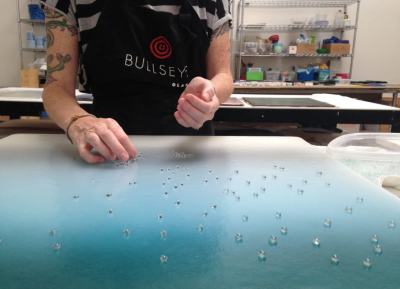 Person wearing a black apron with a "Bullseye Glass" logo printed on it arranged small glass spheres on a blue piece of flat glass.