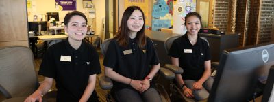 Three smiling youths in black UW branded uniforms sit at a front desk.