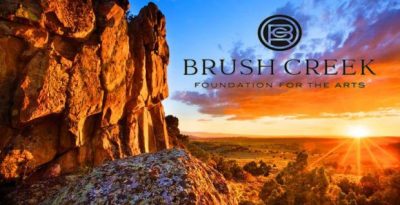 Sunrises over a scrubland desert-scape, a large rock formation in the left side of the frame. Text over the image reads "Brush Creek Foundation for the Arts"