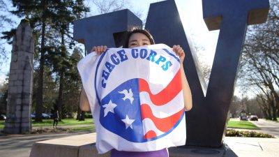 Student stands in front of large metal "W" statue holding a Peace Corps flag in front of their face and torso.