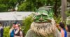 an individual dressed in a troll mask stands in the foreground. blurry parade goers are seen in the background.