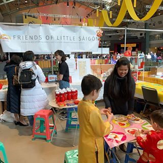 Interior of a large event space full of people, a large banner in the front that reads "Friends of Little Saigon". In the foreground are tables set up with food, and small groups of people eating and chatting.