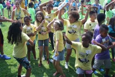 a group of many active youths in a field dancing and jumping around together, all wearing yellow, purple, blue, or green tshirts that read: Miller Summer Day Camp