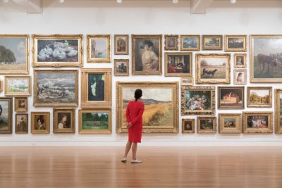 person in a bright red dress stands in front of a gallery wall with multiple framed paintings displayed salon style.