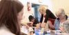 Adults sit at a table working on paintings in a studio setting, while an instructor leans over one student offering guidance.