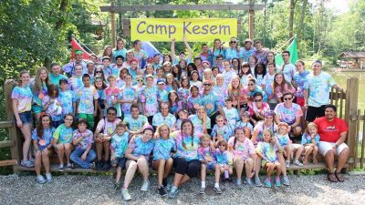 a large group of roughly 30 - 40 individuals stand together in a forested area, with a yellow banner above them reading CAMP KESEM. The group is a mixture of different aged youths who are camp goers and adults who are camp counselors.